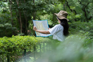 Fototapeta premium Side view of glasses girl holding a map, tourist in the forest - Young woman with white shirt holding and looking at the paper map