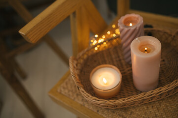 Burning candles on wicker tray in a cozy room with lights on the background.