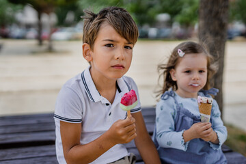 Two children eating ice cream, one preschooler boy and one toddler girl, brother and sister sitting in the park, summer outdoor concept