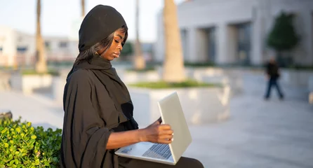 Foto op Canvas Side view of beautiful African female student in abaya with laptop sitting outdoors with her collage in the background. © Bojan