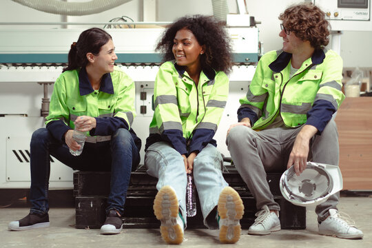 Break time. Group of diverse engineer people sitting together take a break and talking. Young multiracial woman and colleagues technician relax after job in factory. Manufacturing employee lifestyle.