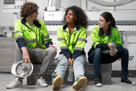 Break time. Group of diverse engineer people sitting together take a break and talking. Young multiracial woman and colleagues technician relax after job in factory. Manufacturing employee lifestyle.