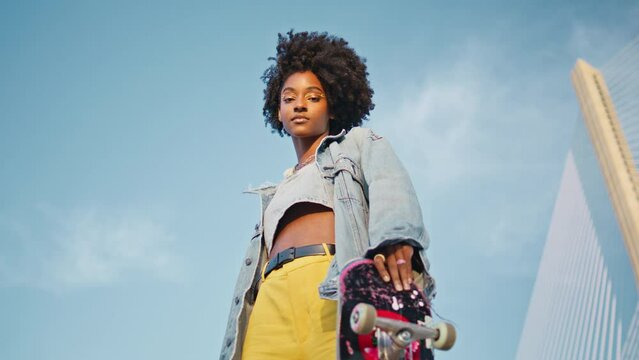 Trendy girl skateboarder looking camera at blue sky. African teenager posing