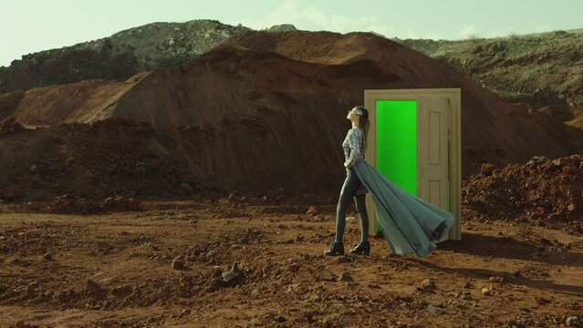 Footage of woman walking on red sand near door with green screen . Young stylish female artist stand near landscape  at day time . Long skirt fabric flowing by wind . Concept of fantasy mystic dreams