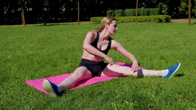 Attractive sporty fit woman doing warm up yoga exercise on fitness mat, stretching leg muscles by touching toe, increasing flexibility and preventing lower back pain while working out outdoors.