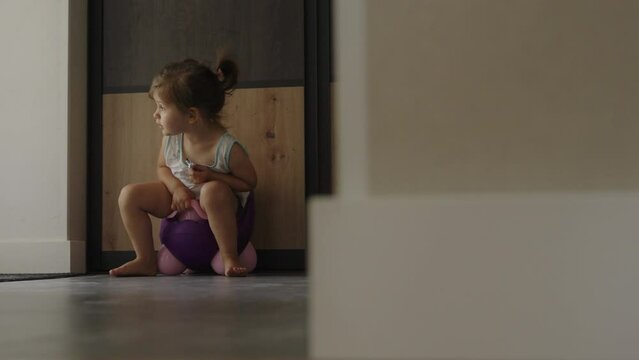 Infant child baby girl toddler sitting on potty, looking away in living room, indoors. Baby hygiene. Playing child.