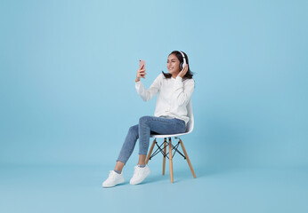 Cheerful young asian girl in wireless headphones sitting in armchair, listening to music, raising her arm, dancing to favorite song, enjoying popular soundtrack isolated on blue background.