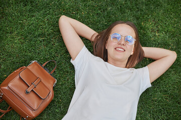 Smiling relaxing brunette woman lying down on grass with peace and tranquility wearing white T-shirt resting in nature smiling happily.