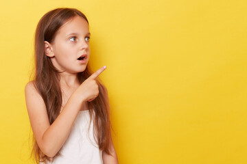 Shocked astonished little girl with ponytails standing isolated over yellow background pointing away at copy space for promotion, advertisement area. - obrazy, fototapety, plakaty