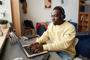 Serious young black man in headphones and yellow sweater sitting at table in students room and using laptop