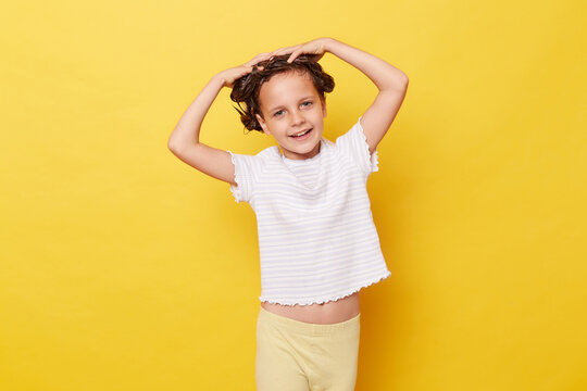 Charming cute little girl with wet hair wearing casual white T-shirt standing isolated over yellow background washing hair with shampoo looking at camera