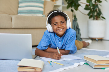 Portrait of happy African-American boy lying on floor and doing hometask while listening to audio in headphones