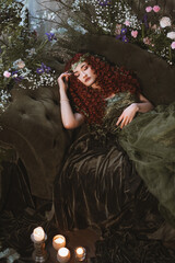 beautiful redhead druid woman on the sofa. young nymph with flowers and candles. fantasy room...