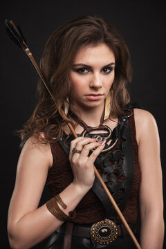 Fantasy fashion warrior woman with an arrow looking at the camera. Medieval archer female studio photography on the black background. Elf lady in boho clothes.