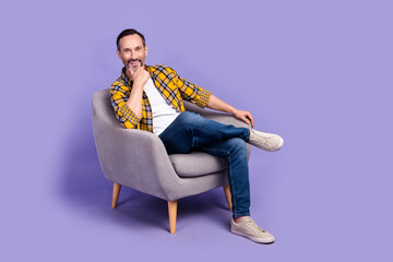 Photo of positive cheerful man wear trendy clothes relax rest chair hand touch chin enjoy comfort isolated on purple color background