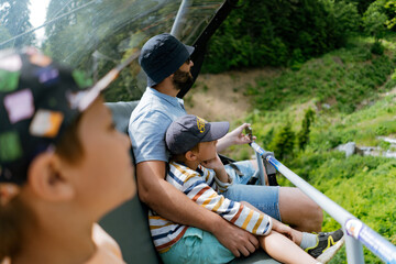 Father and children riding on chairlift in mountains. Modern rope way cabins with people against green valley in mountains © Yulia Raneva