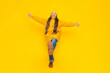 A little girl in full height in an autumn coat, down jackets and hats, rejoices in autumn. The child joyfully raises his leg with his arms outstretched to the sides. Yellow isolated background.