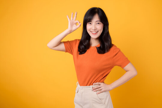 Confident young Asian woman in her 30s wears orange shirt, displaying okay sign on vibrant yellow background. Positive hand gesture concept.
