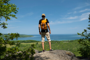 Young man standing on the edge of a rock at lookout viewing panorama of crystal clear turquoise Georgian Bay water and sandy beaches with lush green forest around it. Summer day, clear blue sky. 