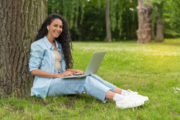 Side view of a young beautiful Arab woman sitting on the grass in a city park typing on a laptop listening to a lesson. Girl preparing for exams outdoors with pleasure. Remote work and learning