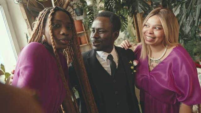 Handheld POV of African American groom with two cheerful bridesmaids recording video of themselves on camera during festive wedding party indoors