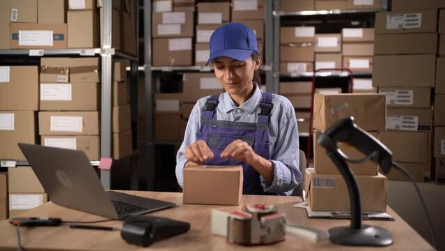 Millennial woman with laptop applying the shipping label to parcel in the warehouse.