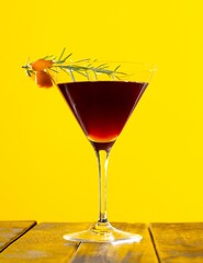 Crystal clear stemmed glass of red cocktail with rosemary on a yellow background