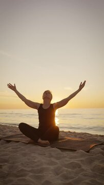 Middle years woman doing breathing exercises raising hands up meditation practice sitting on yoga mat at sunset beach vertical video. Attractive sportswoman making relaxing asana outdoors for
