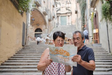 Married tourist couple sightseeing on city street with map - Happy husband and wife enjoying summer vacation together - Tourist lifestyle concept with older woman and man traveling around Girona. - obrazy, fototapety, plakaty