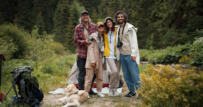 Happy company of tourists: 2 guys and 2 girls posing and looking at the camera in hiking clothes with backpacks against the backdrop of a green forest