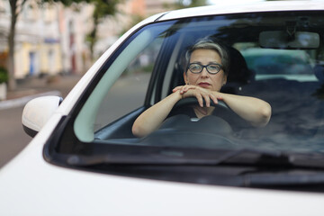 A mature and confident woman drives a white car on a sunny day. She is a successful and happy driver, enjoying her new vehicle.