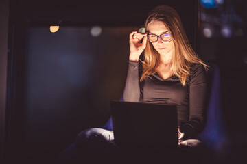 Woman in glasses with a laptop on her lap at night. Face illuminated by a laptop display. - 630825760