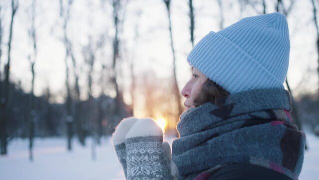 Portrait of a woman in the cold outdoors exhales steam and warms her hands in mittens at sunset, side view