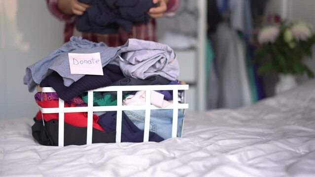 A basket of clothes for donating to Charity shop on bed with blurred woman selecting clothes from her wardrobe. Decluttering Clothes, Sorting, And Cleaning Up. Reuse, second-hand. Conscious consumer.