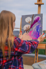 A young girl artist paints a picture with a brush on a canvas that stands on an easel view from behind the back
