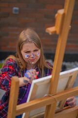 Focused girl artist with paint on her face painting a picture standing on an easel with a paintbrush 