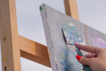 A close-up of a painting spatula applying oil paint to a painting that is standing on an easel in the process of creating a painting