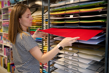 Caucasian woman seller arranges cardboard paper sheets and paper for pastel drawing in shelves in school stationery shop