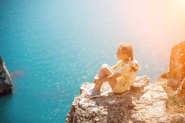 Happy girl perched atop a high rock above the sea, wearing a yellow jumpsuit and braided hair,...