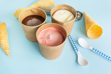 Ice cream in a craft box with different flavors - strawberry, chocolate, vanilla stands on a blue background along with a waffle cone and a checkered spoon. Selective focus, noise.