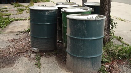 metal barrel drums filled with unknown substance on dirt with sidewalk with pedestrians passing and road behind, blue and green with light color lid