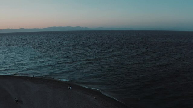 Backing aerial shot of Aegean Sea and pebble beach with sunbeds after sunset, drone footage. Rhodes, Greece.