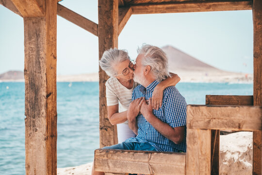 Happy romantic senior couple kissing sitting in front of the sea, two elderly people loving each other and enjoying beach vacation or retirement together