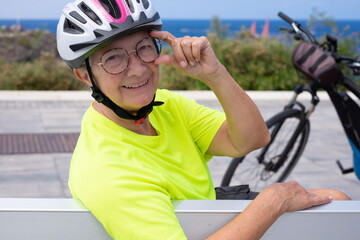 Portrait of relaxed senior cyclist woman sitting in a bench close to her bicycle looking at camera. Elderly carefree lady enjoying freedom, healthy lifestyle and retirement