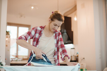 Young pretty woman ironing clean laundry indoors. Young woman ironing clothes. Snow white shirt. Young woman talking on mobile phone standing by ironing board. - Powered by Adobe