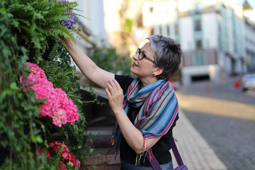 A happy senior woman enjoys her hobby of gardening on a summer day. She is wearing a colorful scarf and holding a purple flower.