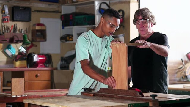 Candid senior carpenter guiding apprentice to use saw machine, slicing piece of wood at workshop