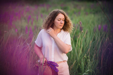 Young woman stands in white shirt in field of purple and pink lupins. Beautiful young woman with curly hair outdoors on a meadow, lupins blossom. Sunset or sunrise, bright evening light