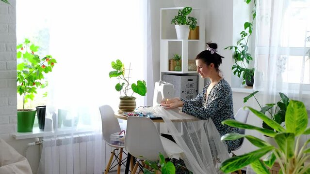 A woman sews tulle on an electric sewing machine in a white modern interior of a house with large windows, house plants. Comfort in the house, a housewife's hobby