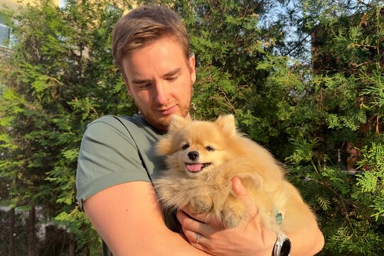 Young man holding on hands his Pomeranian Spitz dog, walking with cute puppy at summer day 